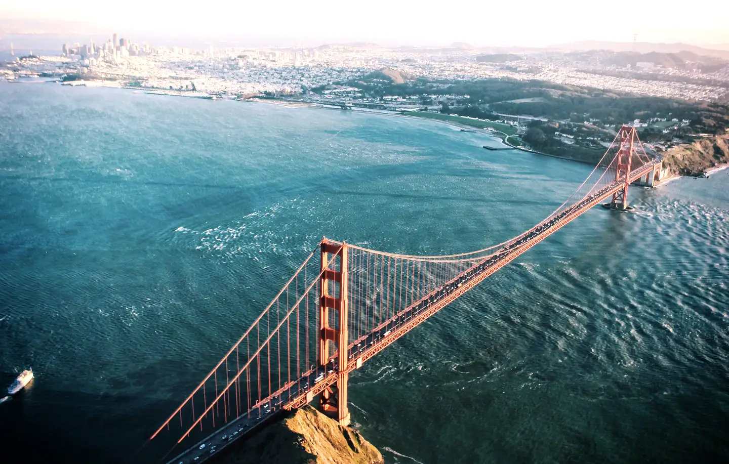 Aerial view of cars crossing the Golden Gate Bridge in warm late-day sun, with San Francisco Bay and the city beyond.