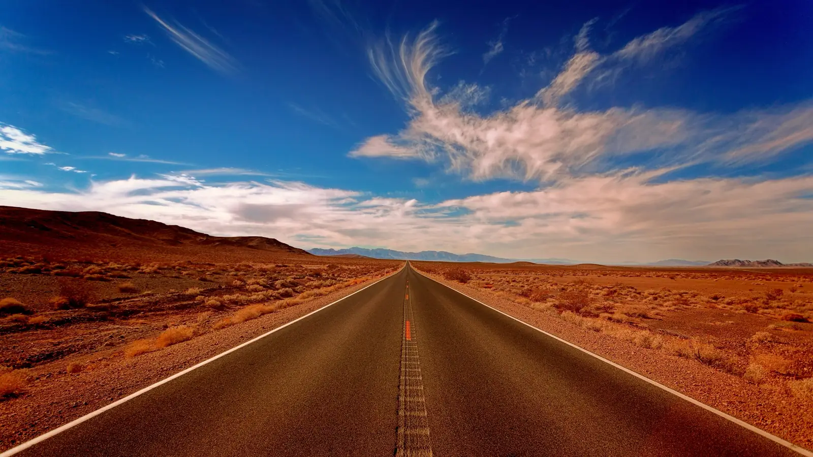A straight desert highway running to the horizon under a wide blue sky with streaks of cloud.